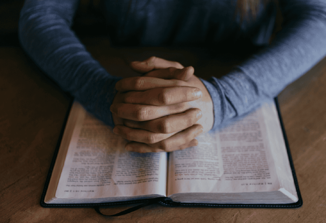 Person praying with hands clasped over an open Bible during a prayer gathering.